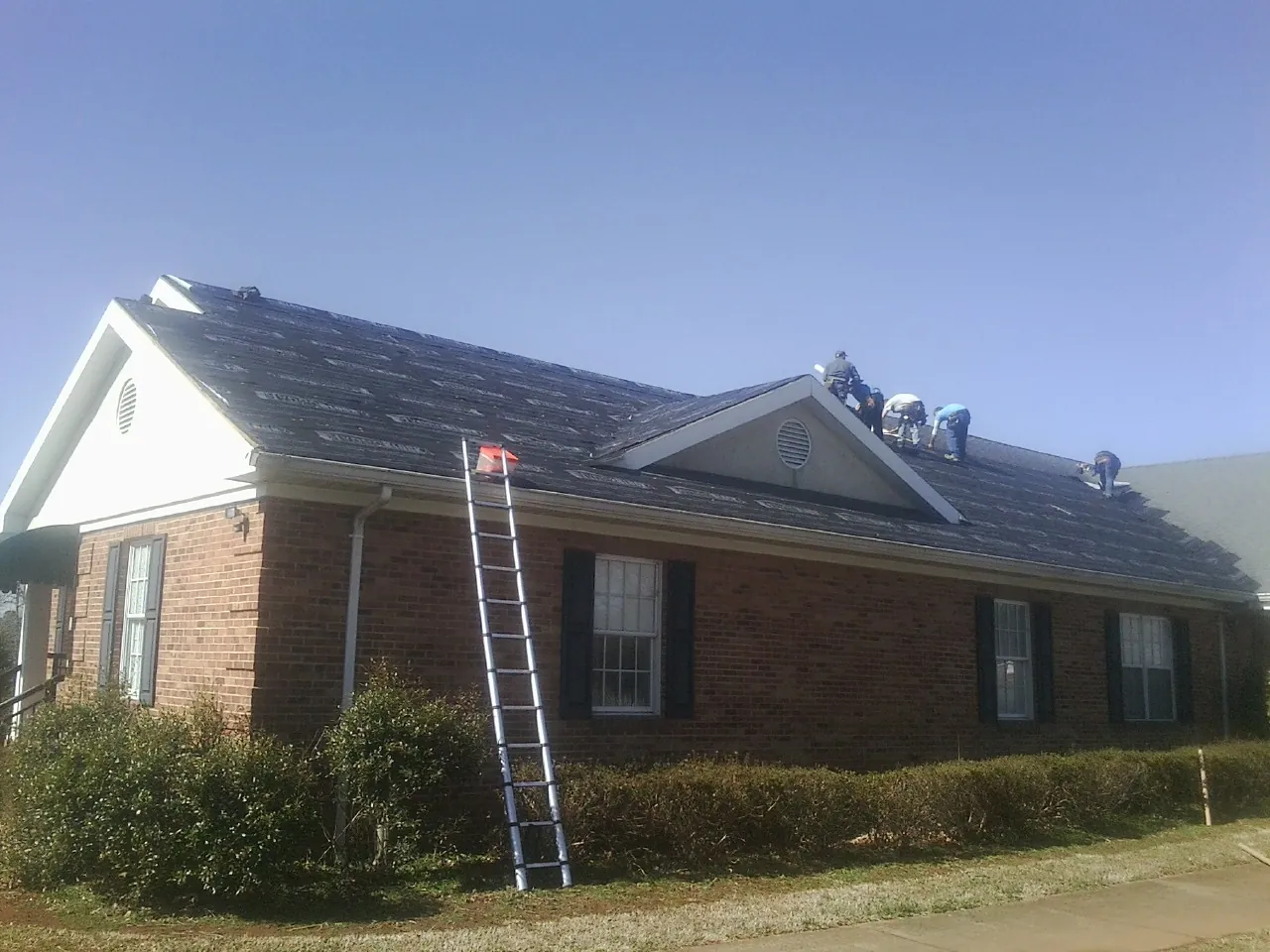 Crew tearing shingles off a brick ranch roof with a ladder set — Metal Roof Repair work by our crew in Monroe