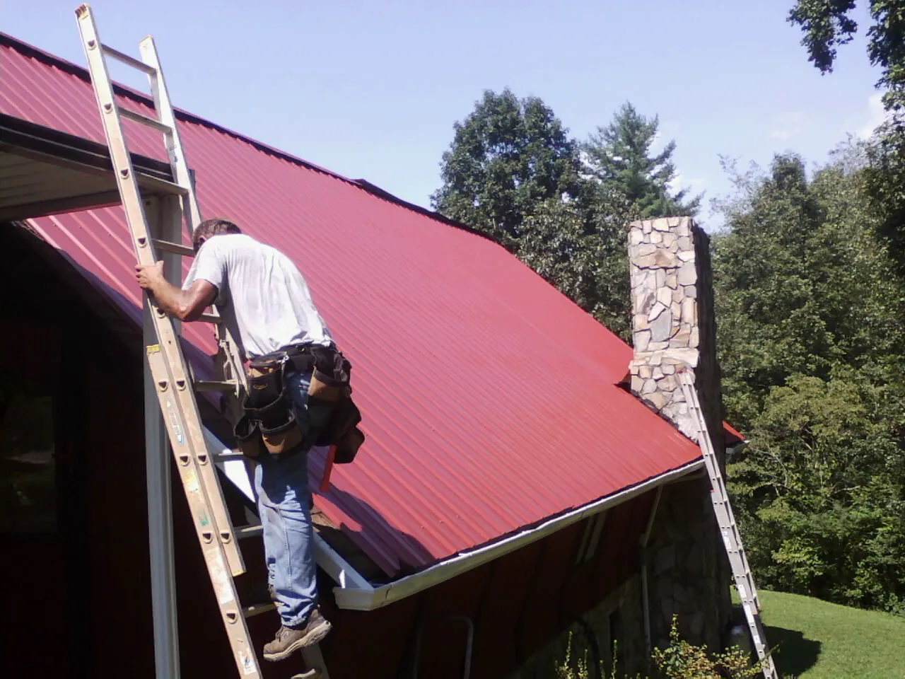 Roofer on a ladder beside a red metal roof and stone chimney — Soffit & Fascia Repair by Roofing Brothers in Monroe